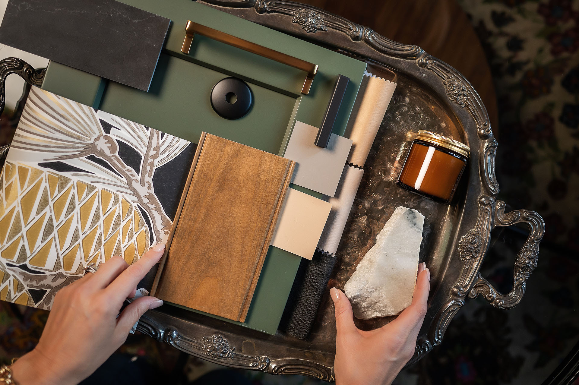 Hands arranging a collection of interior design materials on an ornate silver tray. The materials include fabric swatches, a wooden sample, a green panel with metallic handles, a patterned tile, and a stone piece. A small amber jar is also visible on the tray.